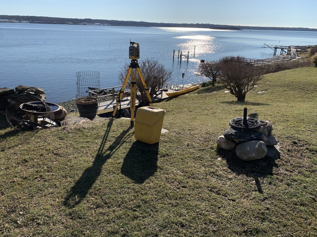 A yellow tripod sits on the edge of a grassy, coastal property in Rhode Island. Docks are seen in the background.