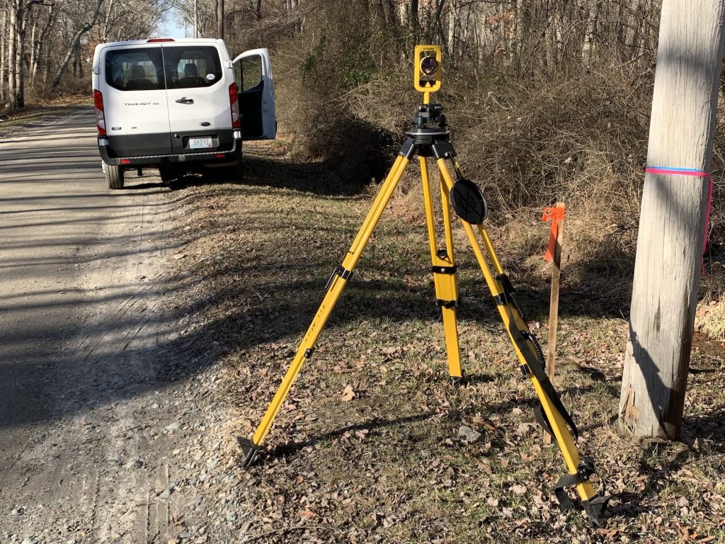 Photo of a yellow surveying tripod on an Able Engineering job site in Rhode Island. The area is partially wooded. A white van is parked on the side of a dirt road.